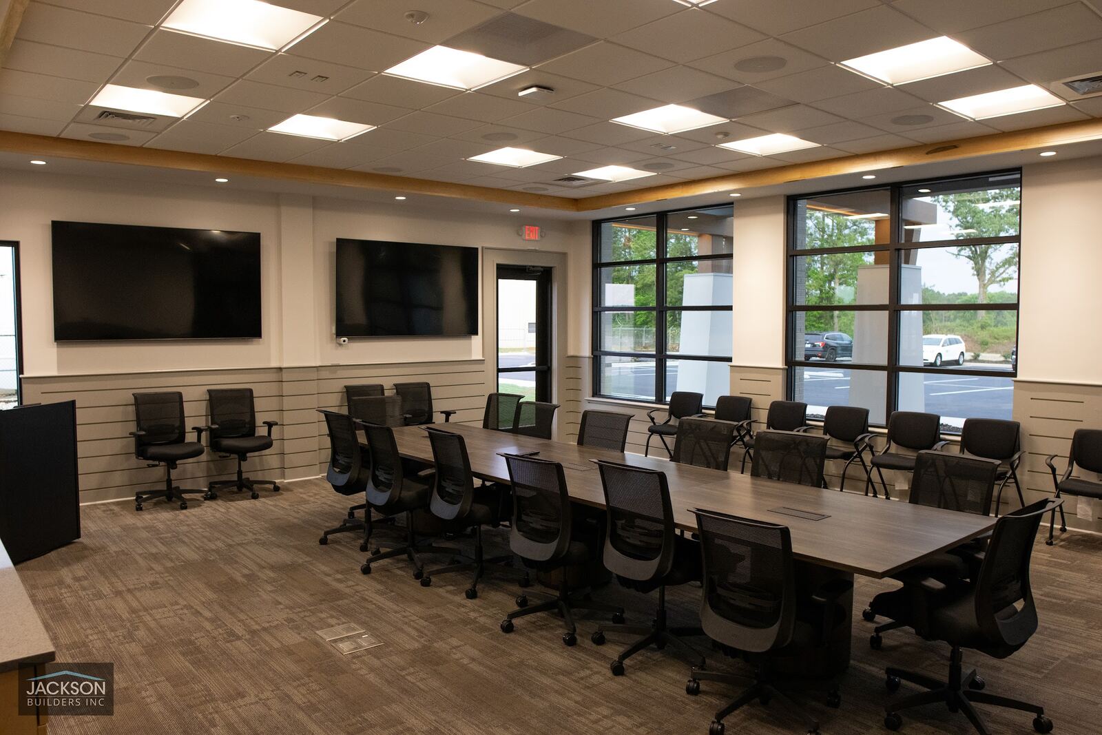 A long conference table lined with black office chairs, screens on the walls and windows to the outside