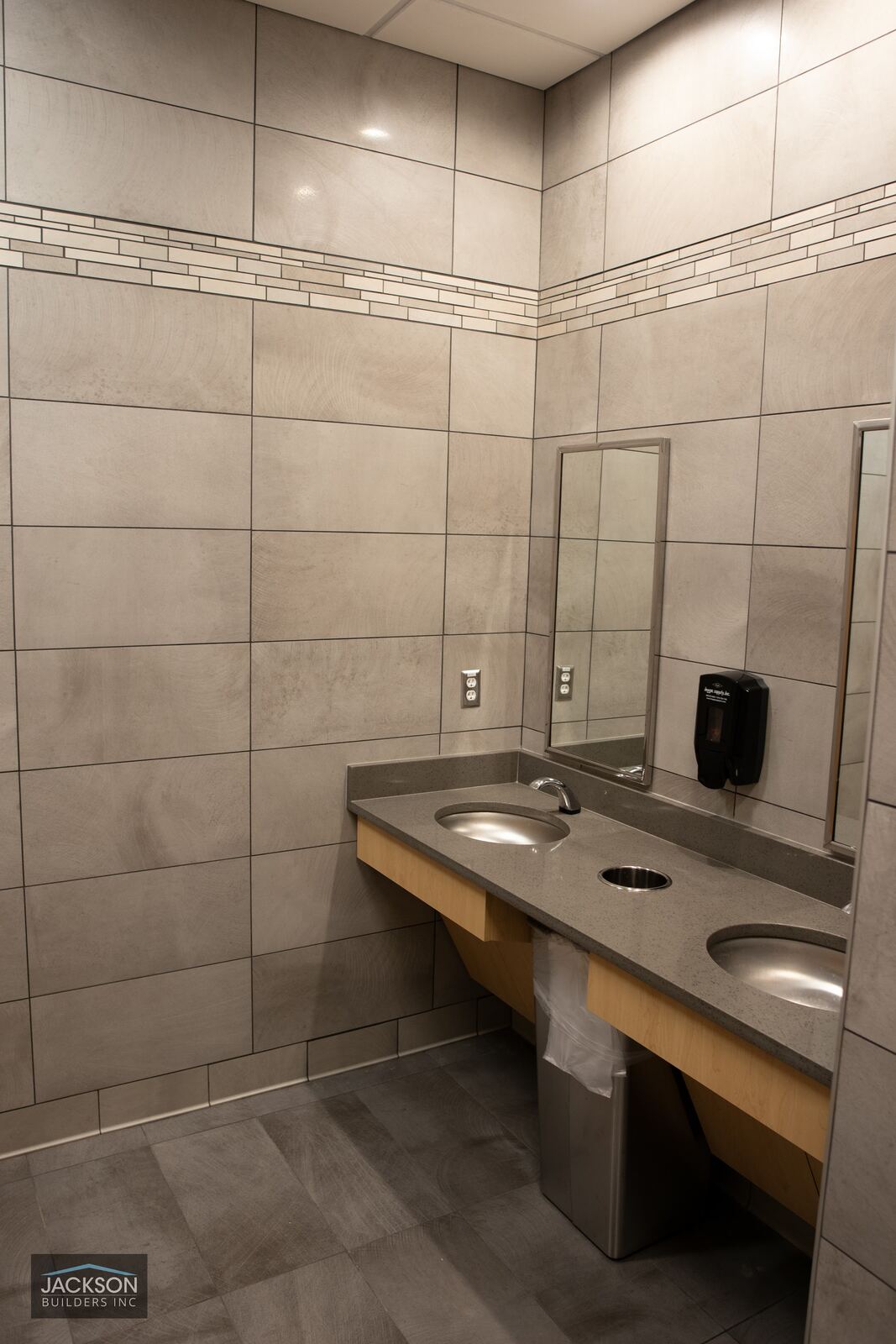 A restroom with large gray tile walls, a wood vanity, and a long mirror